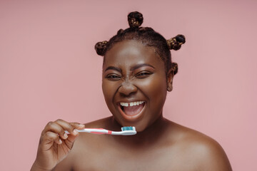 African woman looking in mirror brushing teeth with toothbrush isolated on pink background
