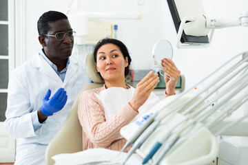 Asian woman patient and african-american man dentist looking at mirror after tooth restoration.