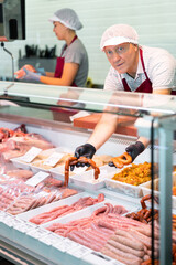 Positive elderly butcher shop salesman offering various traditional Spanish meat products, holding fresh appetizing chorizo and chistorra sausages