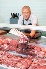 Positive elderly salesman in apron and disposable gloves shows raw beef tomahawk steak . Senior...