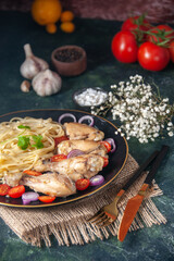 Close up view of pasta meal with chickens and vegetables on a plate on nude towel and cutlery set tomatoes spices on green black mix colors background