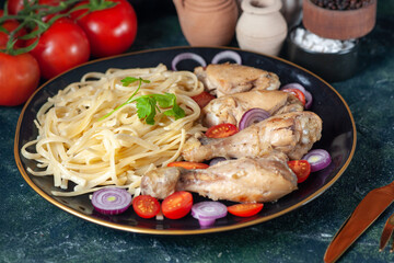 Front close view of raw pastas chickens and vegetables on a plate and cutlery set tomatoes spices cutlery set on green black mix colors background