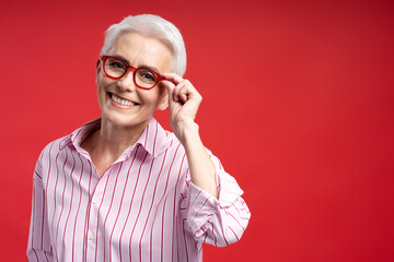 Authentic portrait of smiling senior woman wearing stylish red eyeglasses isolated on red...