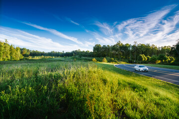 A white car zooms along a curvy road near an open green field in low light