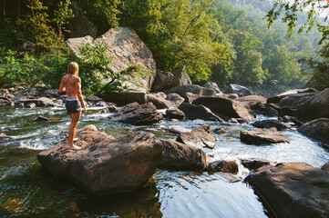 A woman in a swimsuit and bikini top stands on a boulder looking down the river gorge of Little River Canyon National Preserve, Alabama