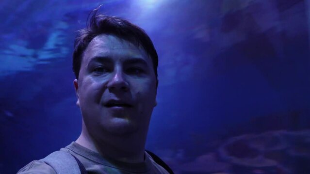 Portrait of male blogger, walks through tunnel of oceanarium with transparent glass roof, blue neon lighting, speaks to camera and shares his impression. Cognitive family leisure aquarium with fish