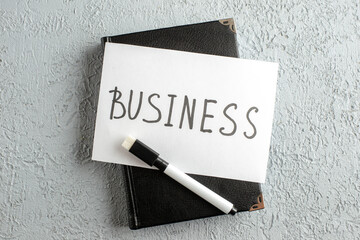 Top view of BUSINESS writing on white sheet and marker on new black closed notebook on gray sand background