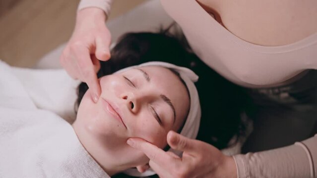 The beautician makes a facial massage. Young woman lies on her back and gets a facelift in a spa salon. Massage techniques for anti-aging.
