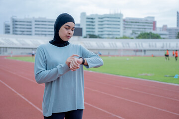 A young asian Muslim woman wearing a black hijab is running and checks progress on a smartwatch with a heart rate monitor at an outdoor stadium in the morning. Modern Muslim woman concept,Muslim sport