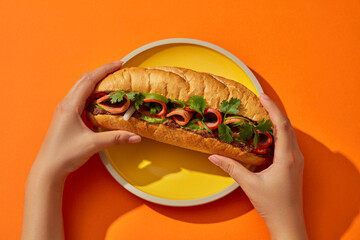 Female hands are holding the ends of the bread and prepare to lift it for breakfast. Bread placed on a round yellow plate on orange background. Top view, creative flat lay