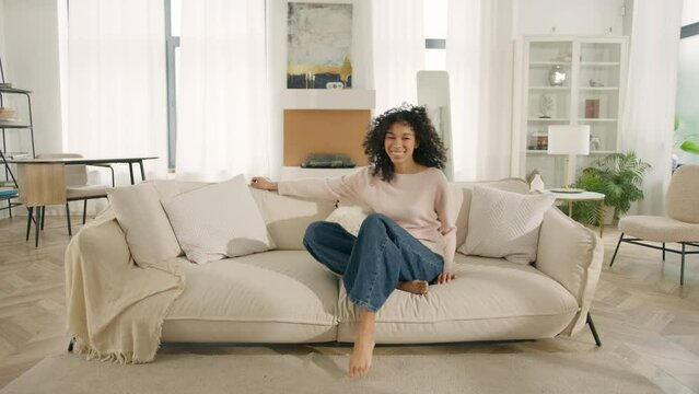 Happy African American woman jumping on modern soft sofa. Curly girl laughing, enjoying rest on cozy couch, feeling overjoyed, celebrating move in day to home, rented flat. Design interior furniture