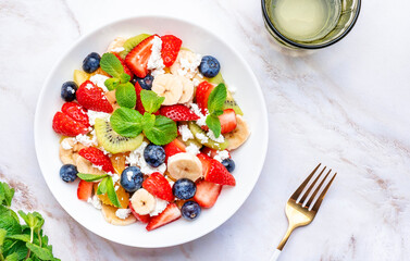 Fresh fruit and berry salad with cottage cheese, strawberries, blueberries, kiwis, oranges and mint leaves with honey dressing, marble table background, top view
