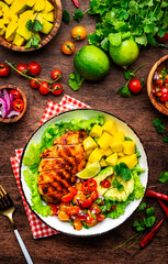 Grilled chicken with side dish salad from avocado, mango, tomato salsa, cilantro, lime and lettuce, rustic wooden table background, top view