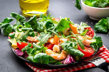 Tasty salad with avocado, shrimps, red tomatoes, cucumber, arugula, lamb lettuce, onion and sesame seeds on black table background. Top view, copy space