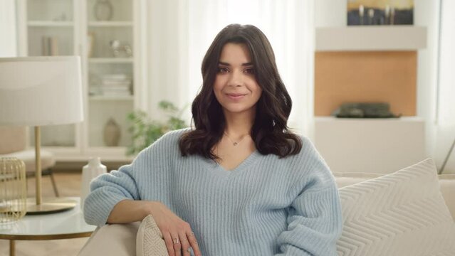 Smiling beautiful hispanic woman looking at camera sitting on sofa alone at home. Happy young latina woman feels satisfied posing for portrait view in living room modern apartment, slow motion people