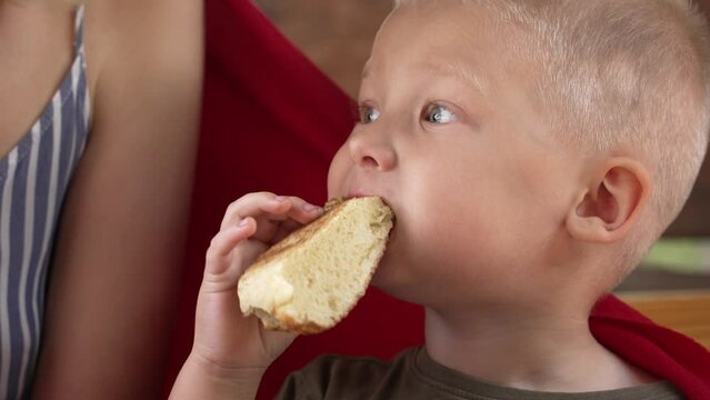 Close-up portrait of a blonde hungry little boy biting a piece of bread while having breakfast with his mother. The son eats in a cafe with his mother. The baby eats nutritious food on his own.