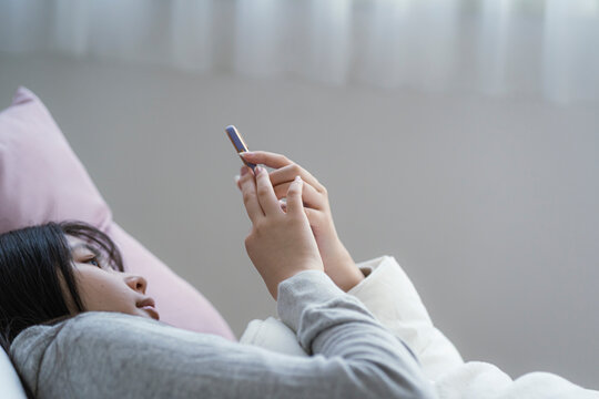 ベッドに横になってスマートフォンを見る若い日本人女性