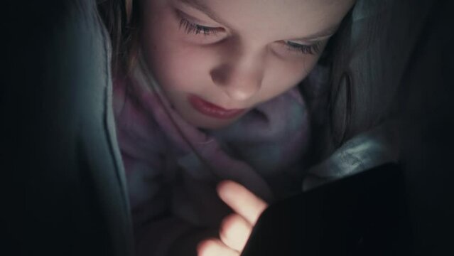 Bedtime phone. Sleepless child. Night online. Curious small girl using gadget under blanket surfing Internet late in bed in dark.