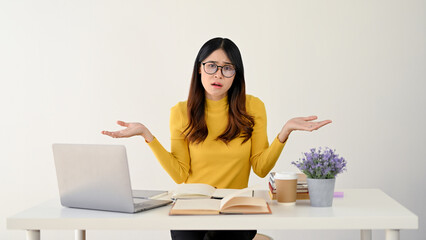 A puzzled Asian female sits at her desk, shrugging her shoulders with hesitation.