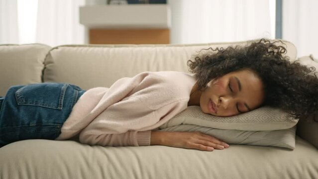 African American girl with curly hair naps turning away on sofa pillow with closed eyes. Nap daydreaming, Night insomnia concept 4K. Tired mixed race woman sleeping on comfortable sofa at modern home