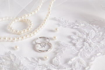 Gentle wedding background. Two white gold engagement rings with a diamond on a white embroidered veil and a pearl necklace. space for text.