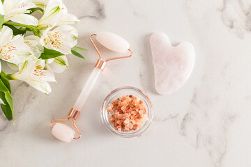 Quartz massage roller and gua sha scraper on a white marble background with astromeria flowers and...