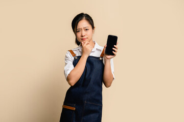 Unhappy young asian barista worker in apron holding smartphone with blank screen. She was wondering...
