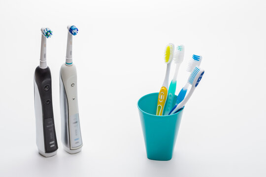 Two Professional Electric Toothbrushes In Front of Four Manual Tooth Brushes in One Cup Over White Background.