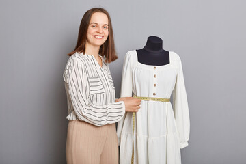 Entrepreneurial clothing venture. Craftsmanship in dressmaking. Beautiful attractive successful woman sewer standing near new dress on mannequin iisolated over gray background.
