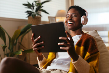 Cheerful young woman wearing wireless headphones watching movie on tablet PC at home