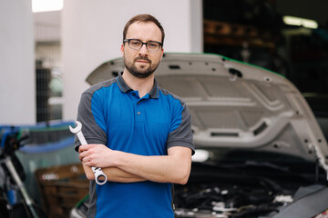 Portrait of handsome male mechanic hold auto repair tool. Man stand in front of car with open hood