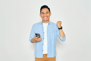Happy young Asian man in casual clothes holding smartphone for trading or chatting and making winning gesture isolated on white background