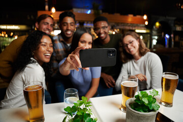 Young multiracial group of friends in casual clothing smiling while taking selfie with mobile phone at bar