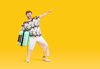 Full body photo of a funny young happy man in sunglasses carrying his blue suitcase is going on summer holiday trip and having fun on a yellow background with copy space. Vacation and travel concept.