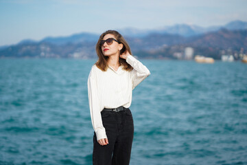 Sea Walk. Young woman wearing sunglasses and casual clothing, posing by sea on Seascape with mountains. Portrait of happy woman walking outdoor in sea. Girl with Shirt and jeans standing on embankmen.