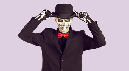 Obraz premium Portrait of man with creepy Halloween skull makeup looking at camera with confident expression. Young man in black suit with bow tie and with skull painted on his face wears hat on lilac background.