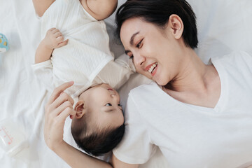 Top view, Beautiful mom and her little baby on bed with happy smile. An Asian mother is happy smile to spend time with her baby.