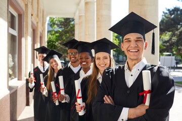 Photo of smiling college graduates standing behind each other. Happy international students in traditional caps and black academic gowns holding diplomas celebrating graduation