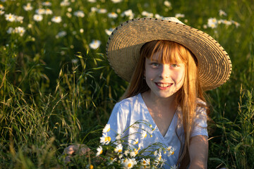 Close up portrait of a happy little cute girl in a blue dress and straw hat on a field with a bouquet of daisies on a sunny summer day at sunset. Summer concept