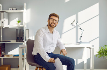 Portrait of a happy businessman at work. Cheerful young handsome business man or office worker in a white shirt, jeans and glasses sitting on a chair by his desk, looking at the camera, and smiling