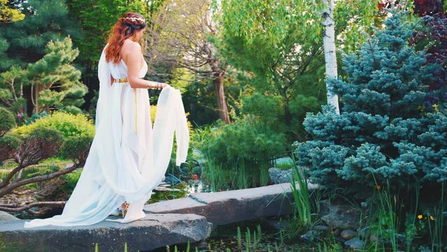 Fantasy happy woman greek goddess walks in spring garden fresh green tree nature day sun light sky. Sexy girl queen antique style white silk dress hem fly in wind. red hair lady bride back rear view