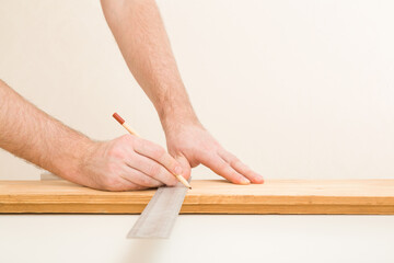 Young adult man hands measuring and marking plank with angle ruler and pencil on white table at light gray wall background. Closeup. Preparing material for repair work of home. Side view.
