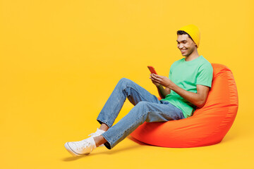 Full body fun young man of African American ethnicity he wears casual clothes green t-shirt hat sit in bag chair hold in hand use mobile cell phone isolated on plain yellow background studio portrait.