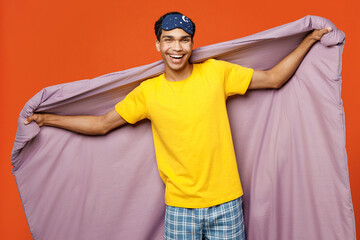Naklejka premium Calm young smiling happy cheerful man wear pyjamas jam sleep eye mask rest relax at home look camera hold blanket duvet isolated on plain orange background studio portrait Good mood night nap concept