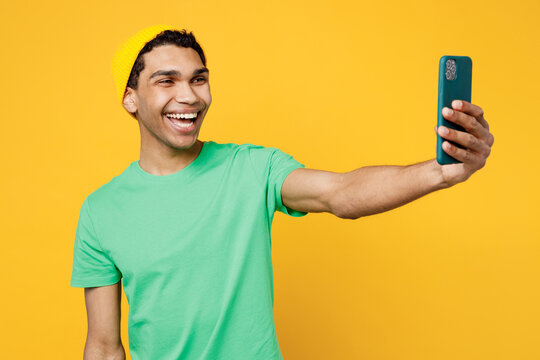 Young man of African American ethnicity wear casual clothes green t-shirt hat doing selfie shot on mobile cell phone post photo on social network isolated on plain yellow background studio portrait.