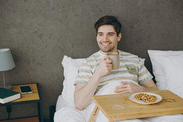 Young man wear casual clothes t-shirt pajama lying in bed eat cookies drink coffee tea have breakfast rest relax spend time in bedroom home in own room hotel wake up dream be in reverie good mood day.