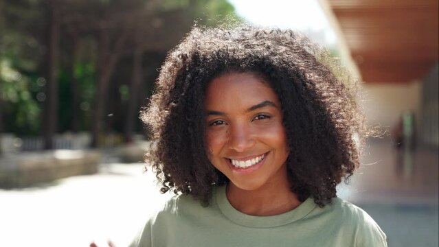 Young happy cute pretty young positive beautiful African American ethnic woman model with curly hair and dental smile looking at camera in city street outdoors on sunny day, close up face portrait.