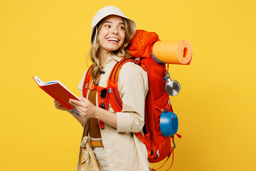 Young woman carry backpack with stuff mat read book diary look aside on area isolated on plain yellow background Tourist leads active lifestyle walk on spare time Hiking trek rest travel trip concept