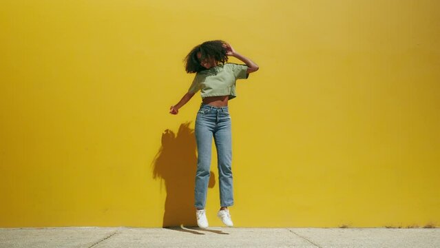 Young free funny excited happy African American ethnic woman wearing sunglasses jumping dancing having fun at yellow wall background feeling joy on sunny summer day outdoors.