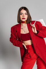 consumerism, bold makeup, tattooed young woman with brunette short hair and nose piercing holding shopping bag and standing on grey background, youth culture, fashionable outfit, red suit